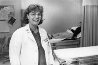 Vanderbilt FNP faculty Charlotte Covington, wearing a white coat, stands in a simulated hospital setting with a patient resting in the background. the photo is black and white, and circa 1980s.