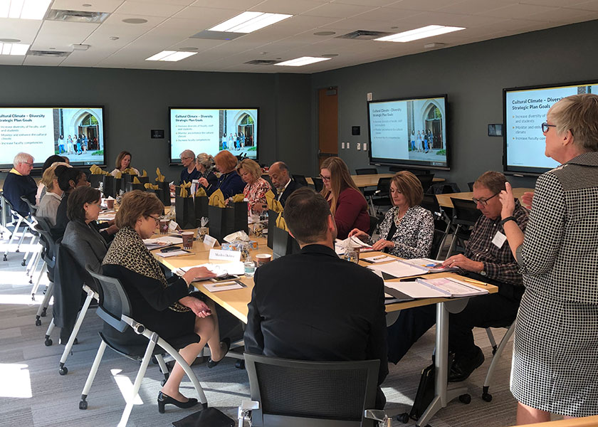 A group of people sit on four sides of a long table and listen to Senior Associate Dean Mavis Schorn talk. The room is modern and has large digital displays on all the walls.