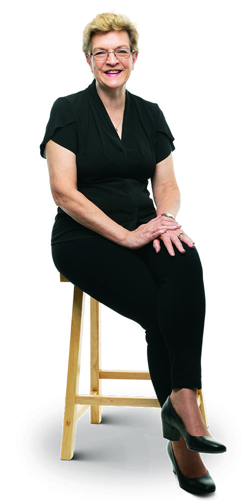 Nurse practitioner Kay Davis in black trousers and blouse, seated on a stool.