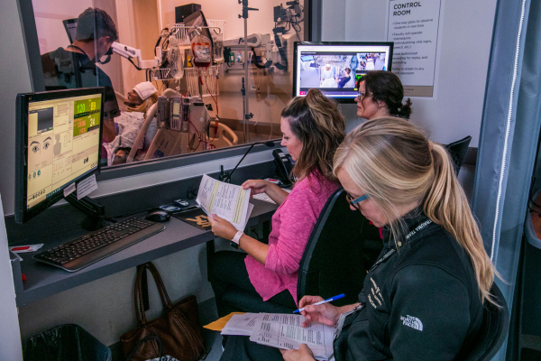 People on one sided glass watch students work with a patient in the simulation lab.