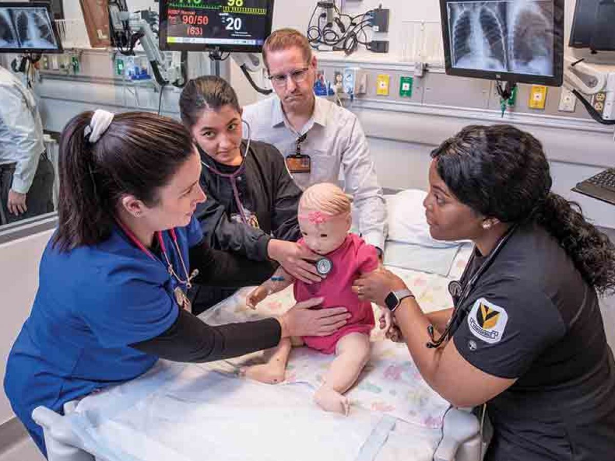 2 nursing students in black scrubs perform a wellness check on a child mannequin as instructor and Sim Lab staffer look on