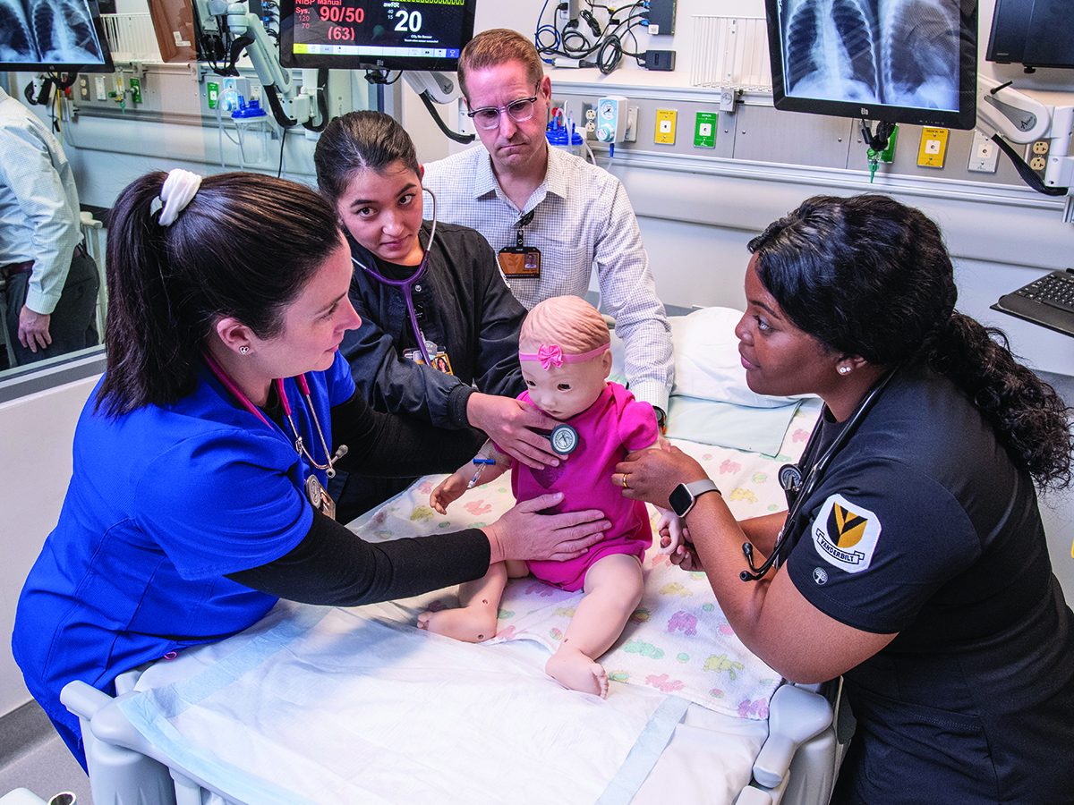 2 nursing students in black scrubs perform a wellness check on a child mannequin as instructor and Sim Lab staffer look on