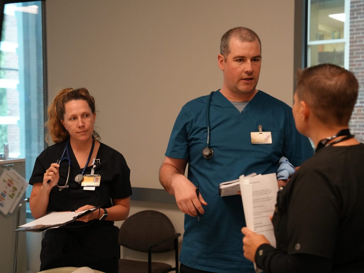 A male student speaks with an emergency provider while another student looks on during a simulation